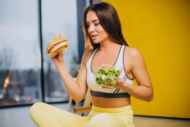 Una mujer con una hamburguesa y una ensalada en la mano. / Freepik.