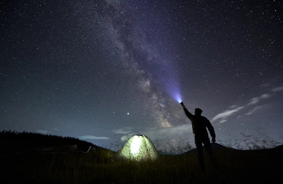 Back view of male traveler illuminating beautiful starry sky. Man hiker shining flashlight into night starry sky with Milky Way near illuminated camp tent. Concept of travelling and night camping.