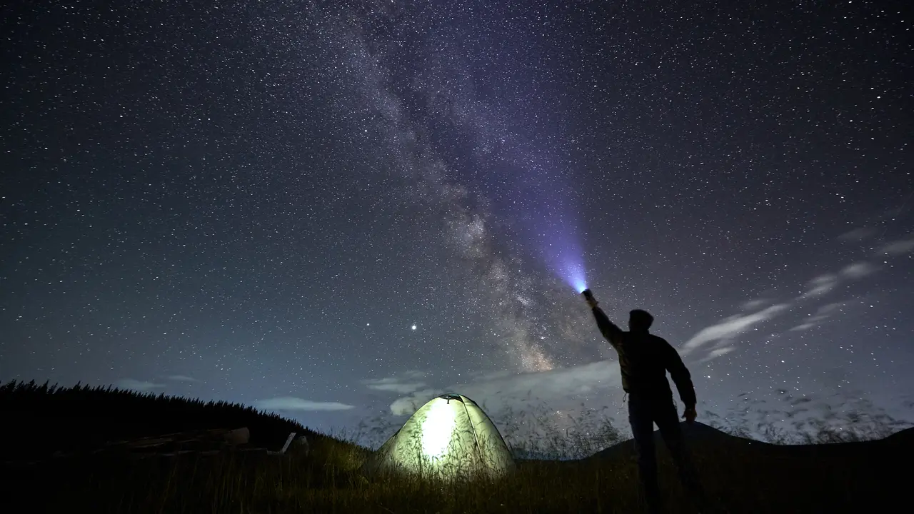 Back view of male traveler illuminating beautiful starry sky. Man hiker shining flashlight into night starry sky with Milky Way near illuminated camp tent. Concept of travelling and night camping.