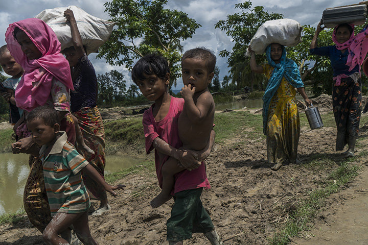 A Rohingya boy carries his baby brother as he crosses into Whaikhyang, Bangladesh with his family. ; As hundreds of thousands of Rohingya sought safety in overstretched camps and settlements in Bangladesh in late-August/early-September 2017, officials warned of a deepening humanitarian crisis. Whole families, young mothers and unaccompanied minors were among those fleeing for their lives since fighting reignited in Myanmar. They came by boat or walked barefoot for days, wading through vast rice fields. They left most of their possessions behind. Large groups crossed into the Ukhiya and Teknaf areas of south-eastern Bangladesh bordering Myanmar. Many were hungry, in poor physical condition and in need of life-saving support. They congregated at long-established camps Kutupalong and Nayapara, as well as makeshift sites and local villages that have hosted several hundred thousand Rohingya since the mass displacement of the early 1990s. “Every available space is occupied,” reported Mohammad Abul Kalam, the Refugee Relief and Repatriation Commissioner in Cox’s Bazar.